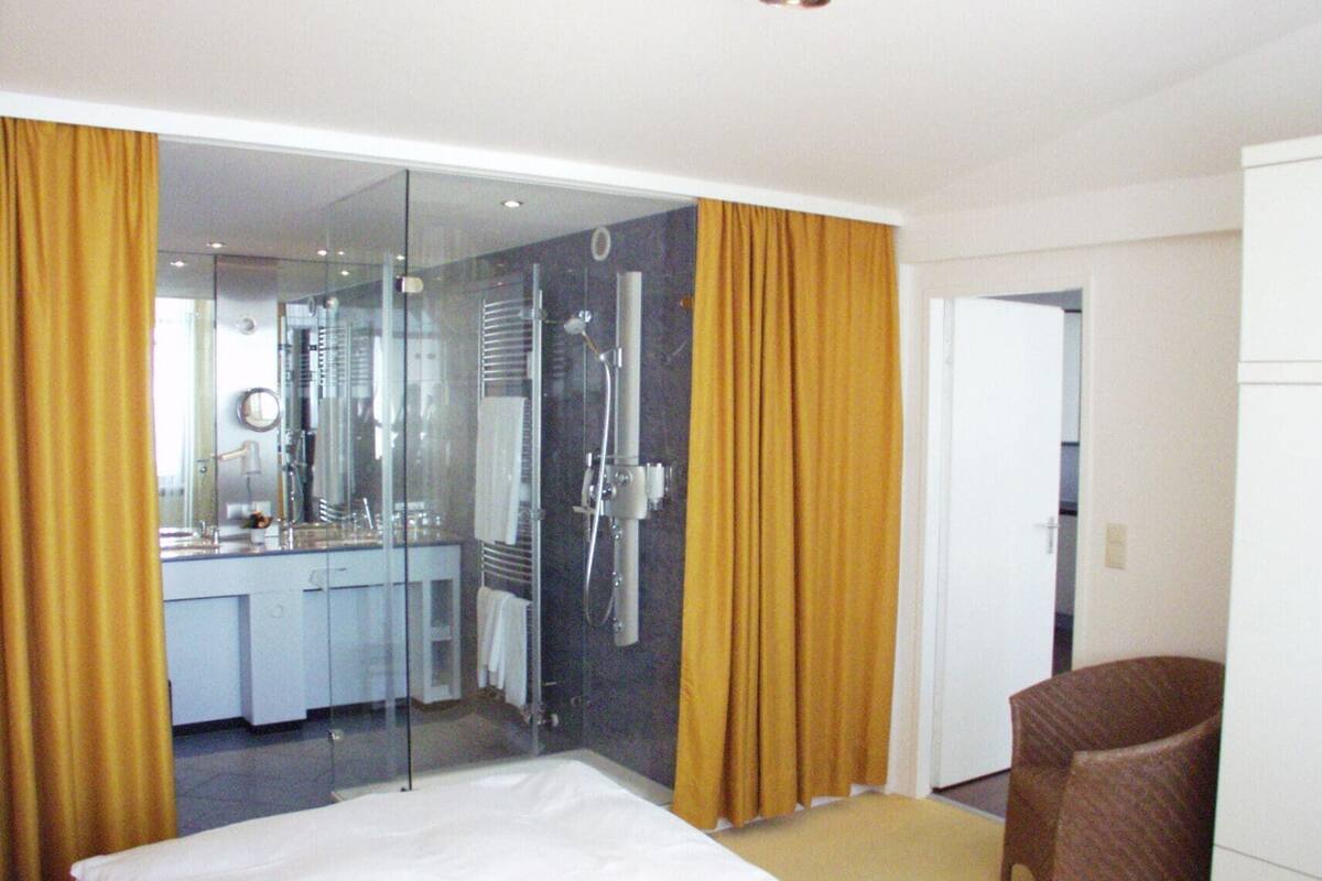A hotel room with a bed, a bathroom with a glass shower enclosure, a mirror, and a chair.