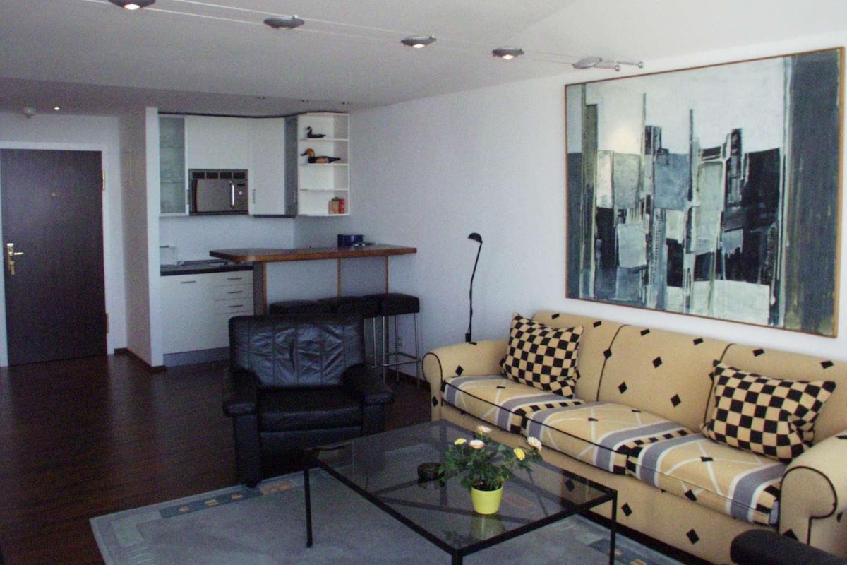 A living room with a sofa, a coffee table with a plant, a kitchen area with a microwave, and a large mirror on the wall.