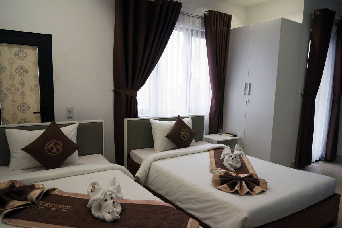 Habitación de hotel con dos camas, cada una con una almohada y una toalla. Hay una mesita de noche con un vaso y un libro. La habitación tiene una ventana con cortinas y una puerta con manija.