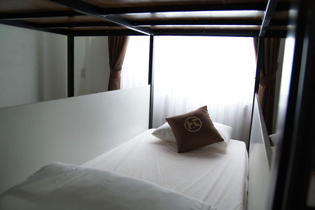 Una cama individual con ropa blanca y una almohada marrón con un logotipo, una ventana con cortinas y una estructura de techo metálico.