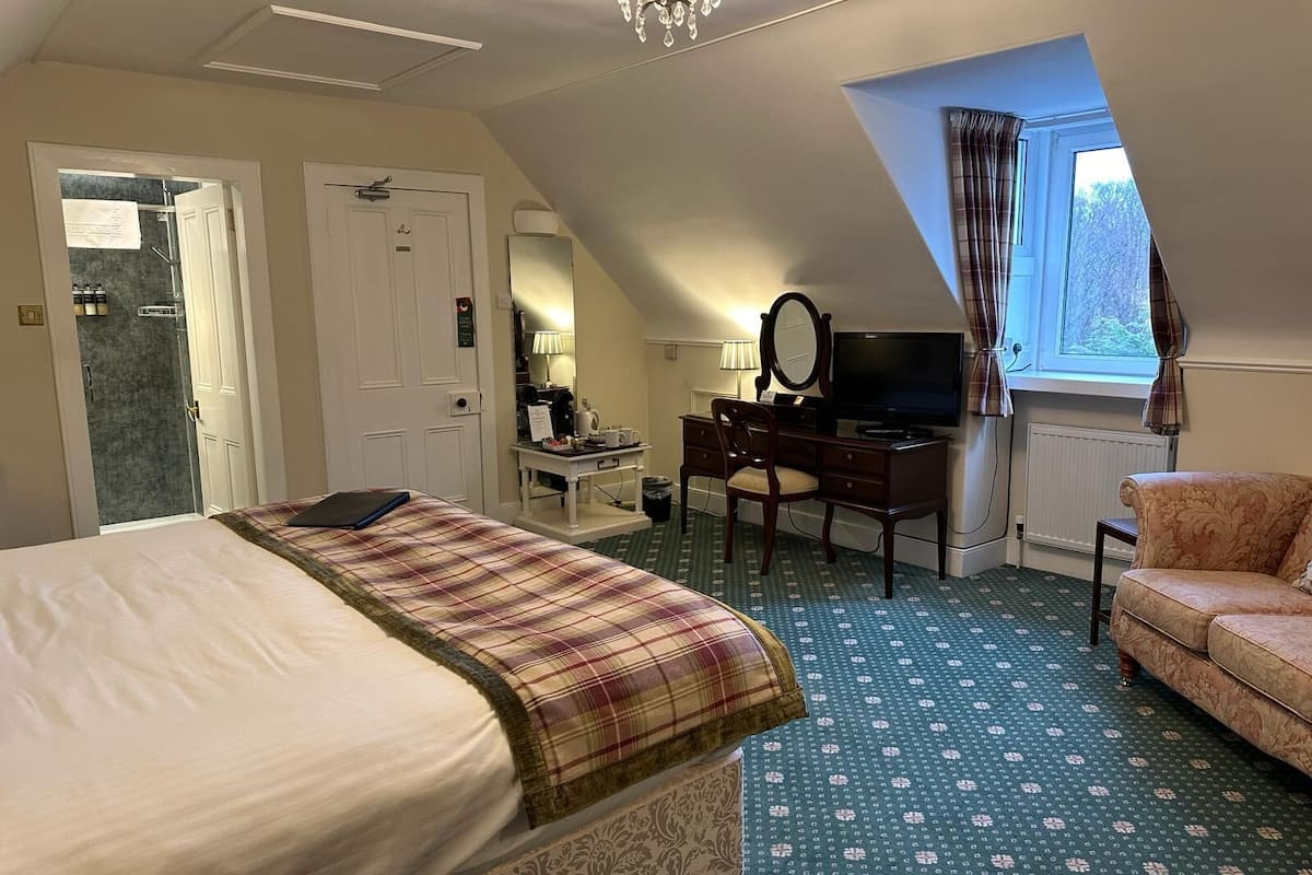 A hotel room with a bed, a desk with a computer, a chair, a sofa, and a window with curtains.