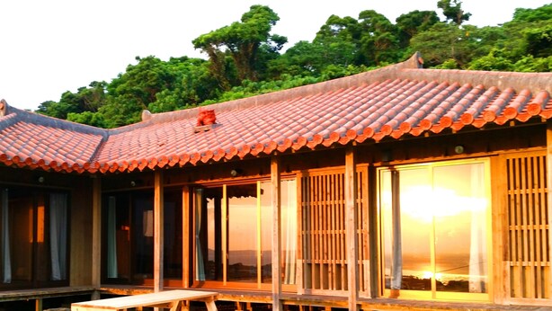 A traditional wooden house with a tiled roof, large sliding glass doors, and a wooden table in the yard. - Churamui Terrace (Motobu)