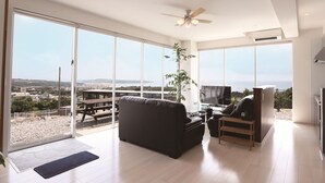 A spacious living room with a large sliding glass door leading to a balcony, a ceiling fan, a flat-screen TV, and a dining area with a table and benches. - Churamui Terrace (Motobu)