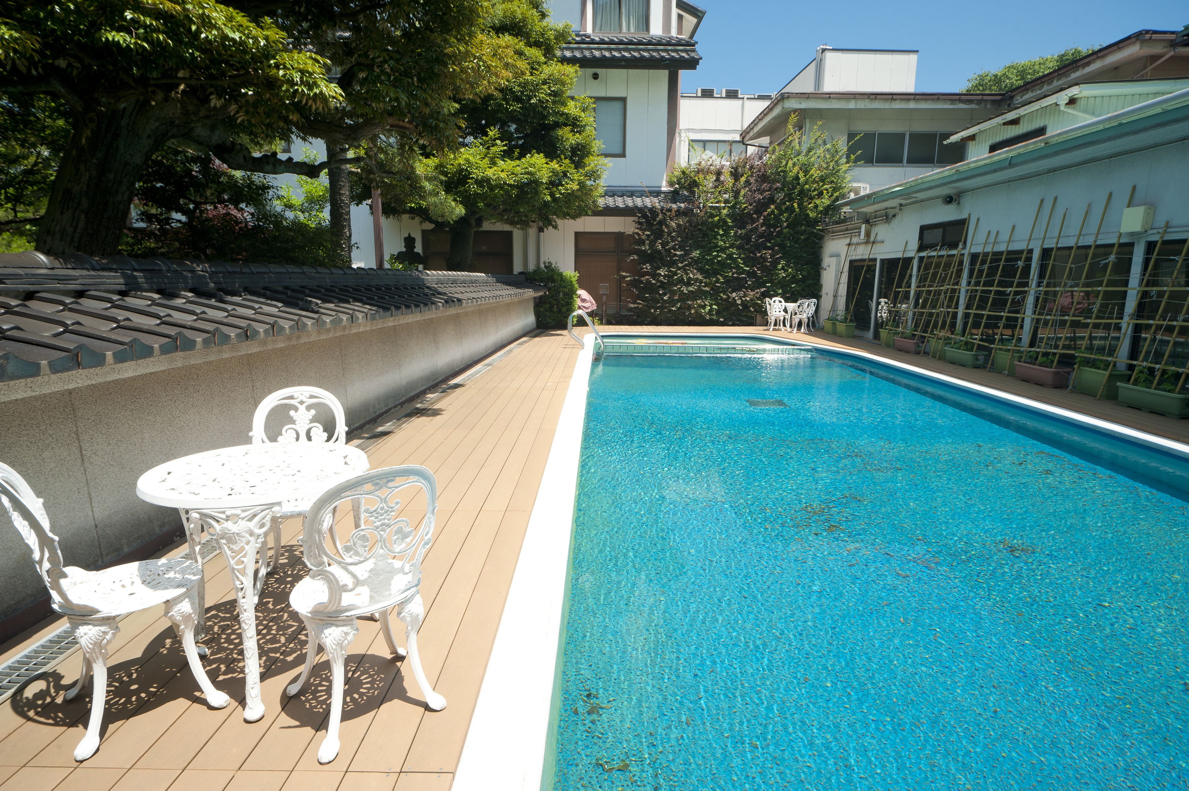 A swimming pool with a tiled deck, surrounded by potted plants and a traditional building in the background.