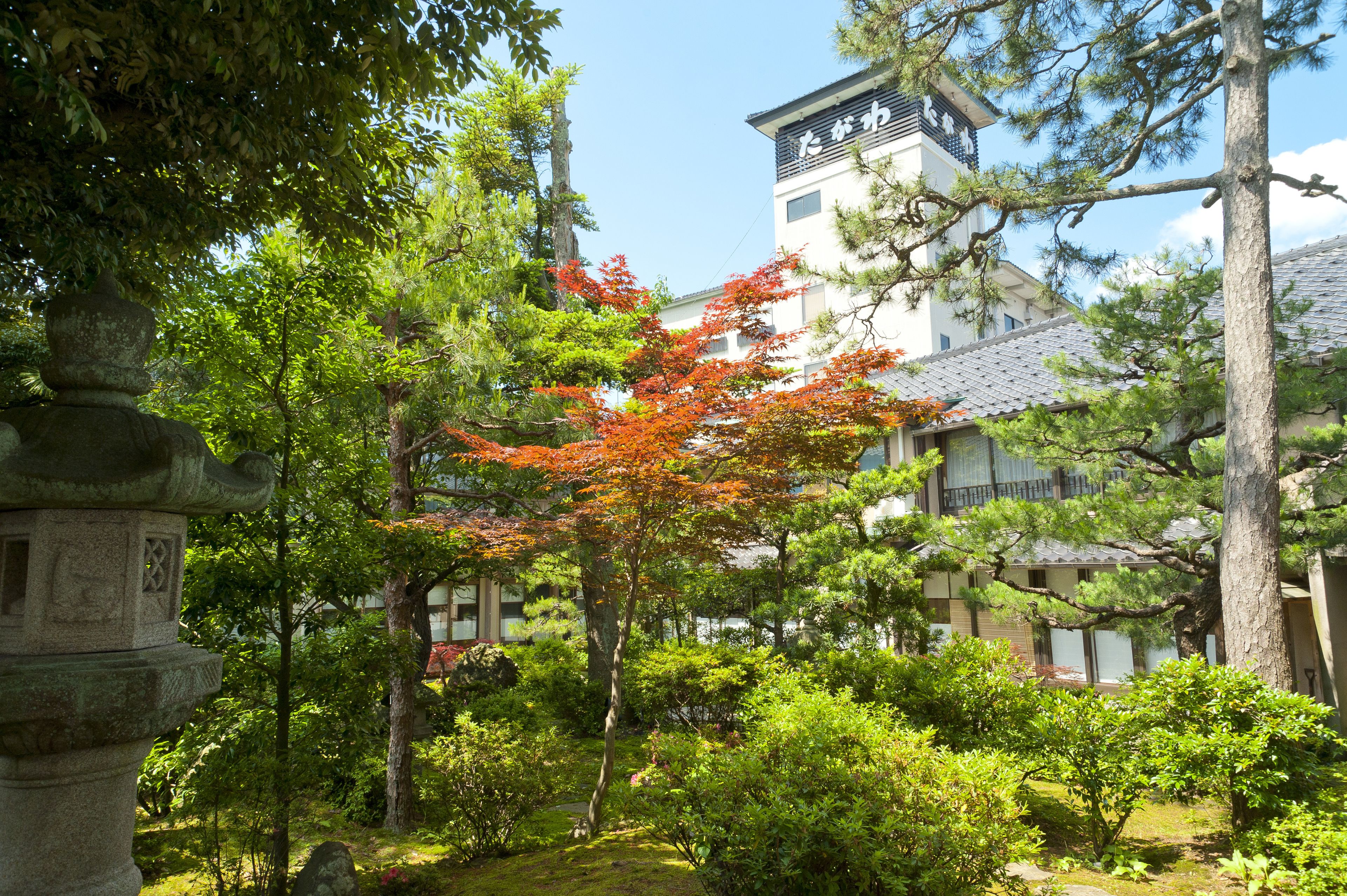 A traditional Japanese building with a stone lantern, surrounded by lush greenery and autumn-colored foliage.