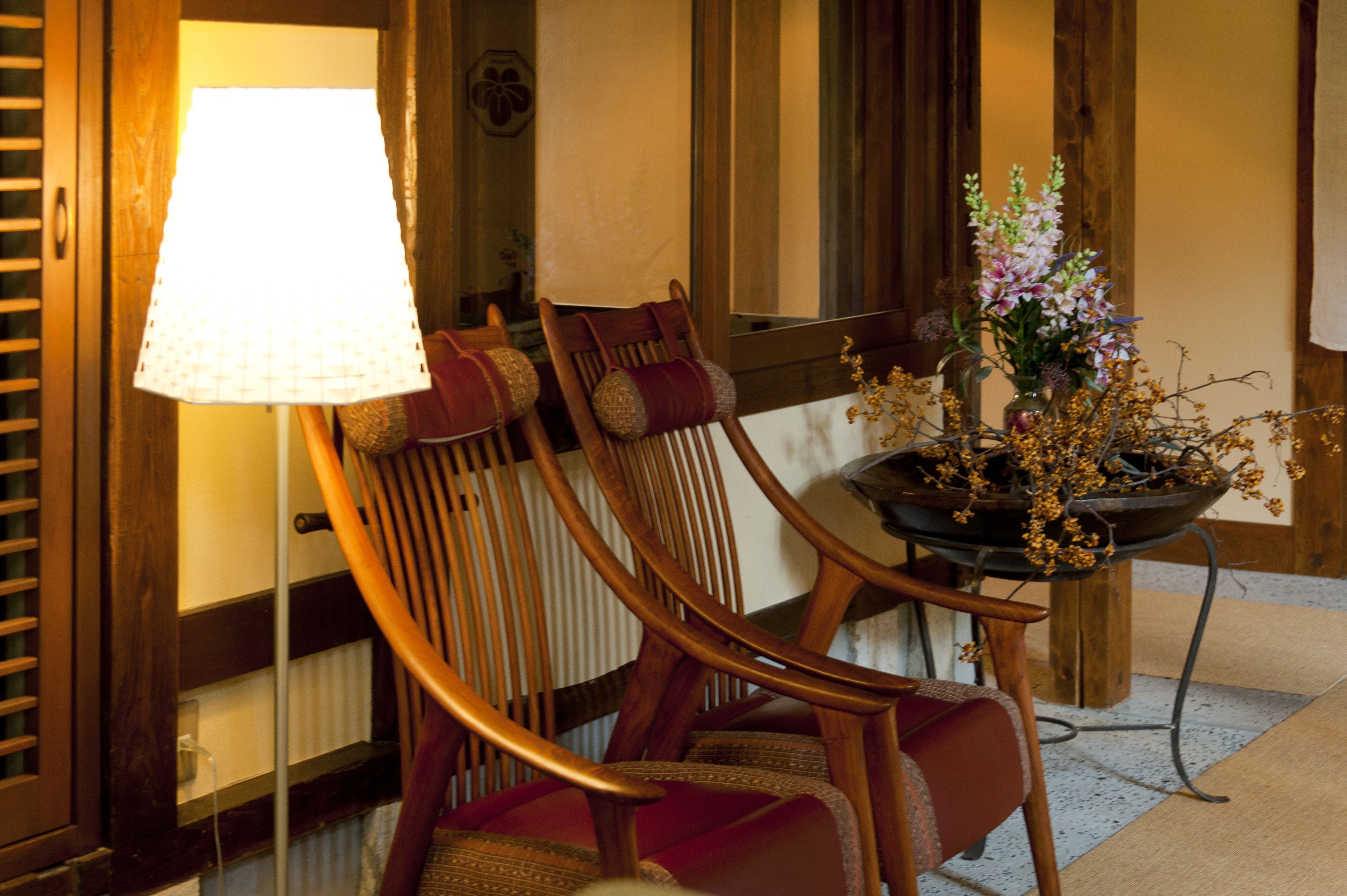 A wooden chair with a cushion, a table with a vase of flowers, and a lamp.