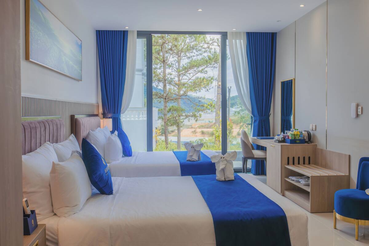 A hotel room with two beds, a desk, a chair, and a view of the outdoors.