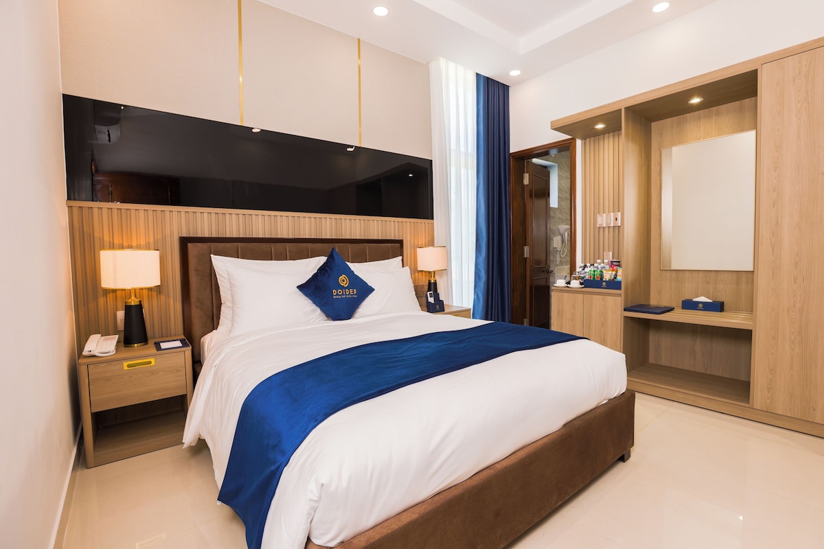 Una habitación de hotel con una cama grande, cabecero de madera, mesitas de noche con lámparas, un armario y un espejo.