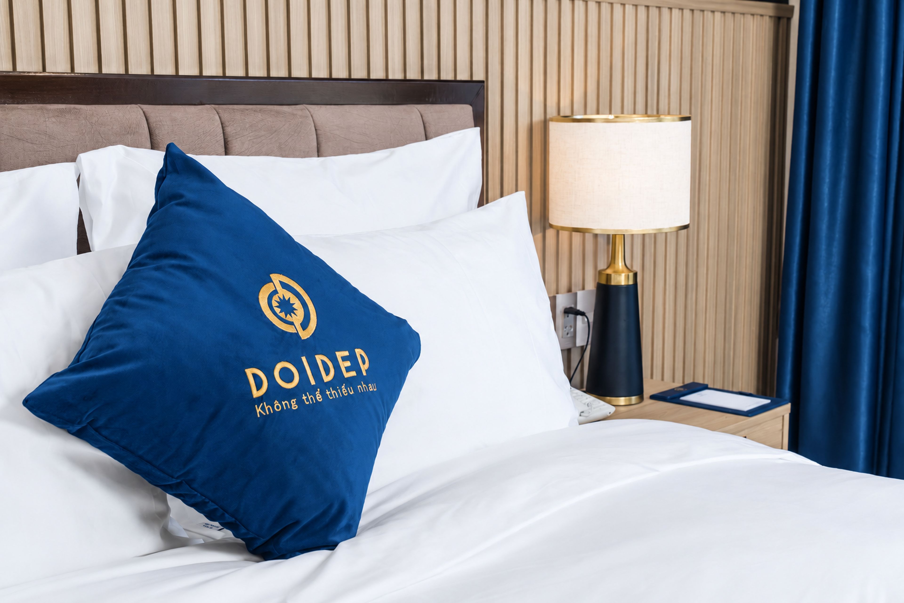 Habitación de hotel con cama, ropa de cama blanca, almohada azul con un logotipo, lámpara de noche y mesita.