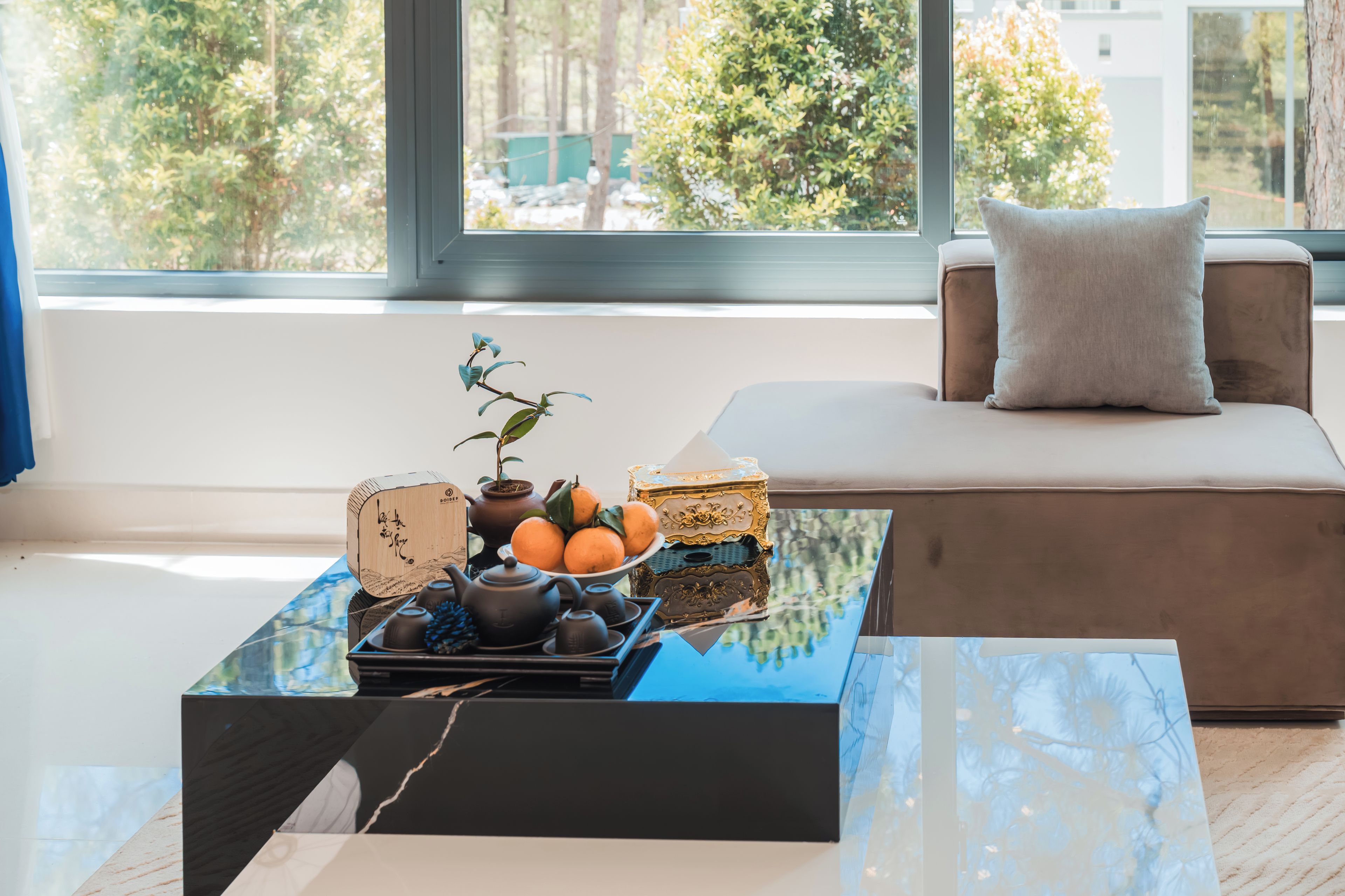 A modern living room with a sofa, a coffee table with a plant and fruit, and a view of trees through a large window.