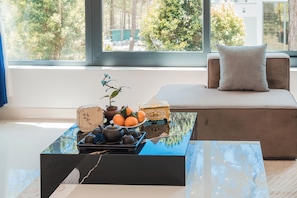 A modern living room with a sofa, a coffee table with a plant and fruit, and a view of trees through a large window.