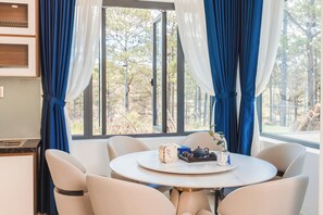 Comedor con una mesa redonda, sillas blancas y vista a árboles a través de amplios ventanales.