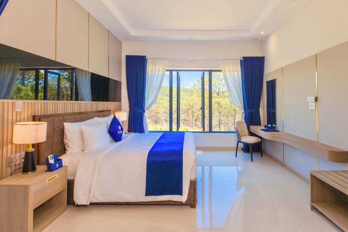 A hotel room with a bed, bedside table, TV, desk, chair, and large windows.