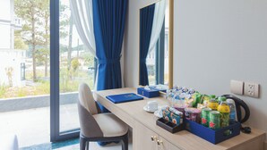 Habitación de hotel con escritorio, silla, espejo, ventana con cortinas y varios objetos sobre el escritorio, incluyendo un cuaderno, una taza y una caja de aperitivos.