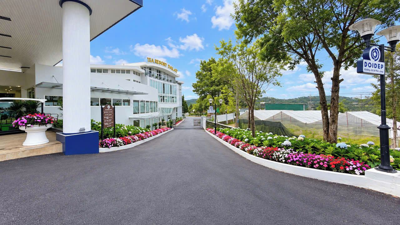 Un edificio moderno con un cartel que dice 'TEA RESORT DALAT' y un sendero bordeado de flores coloridas que lo conduce.