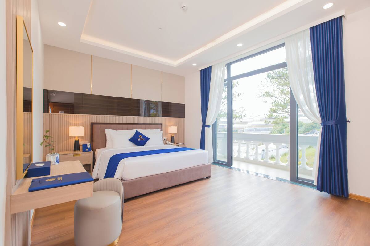 A modern hotel room with a large bed, a desk, a chair, and a balcony with curtains.