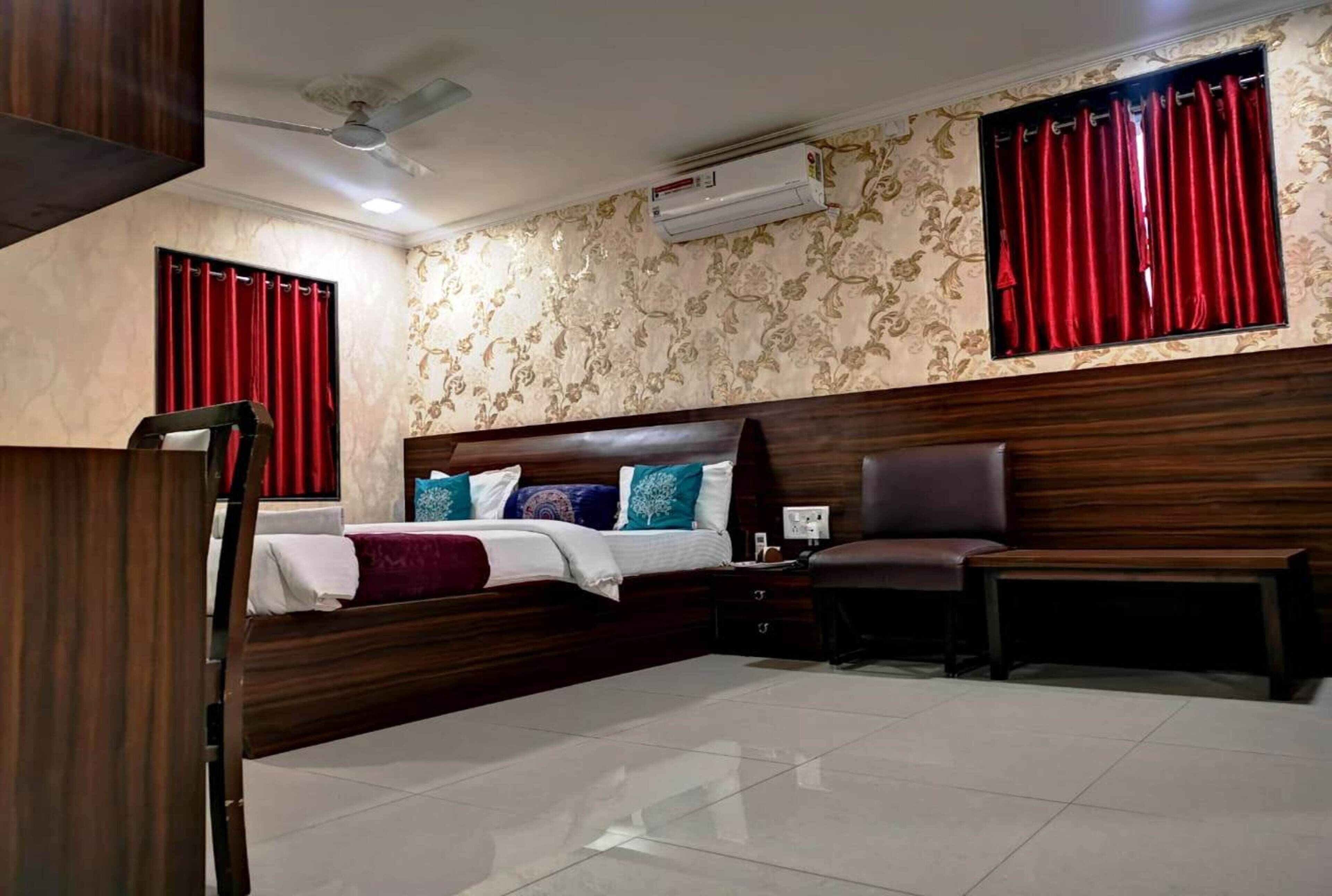 A hotel room with a wooden headboard, a bed with multiple pillows, a bench, a nightstand, and a window with red curtains.