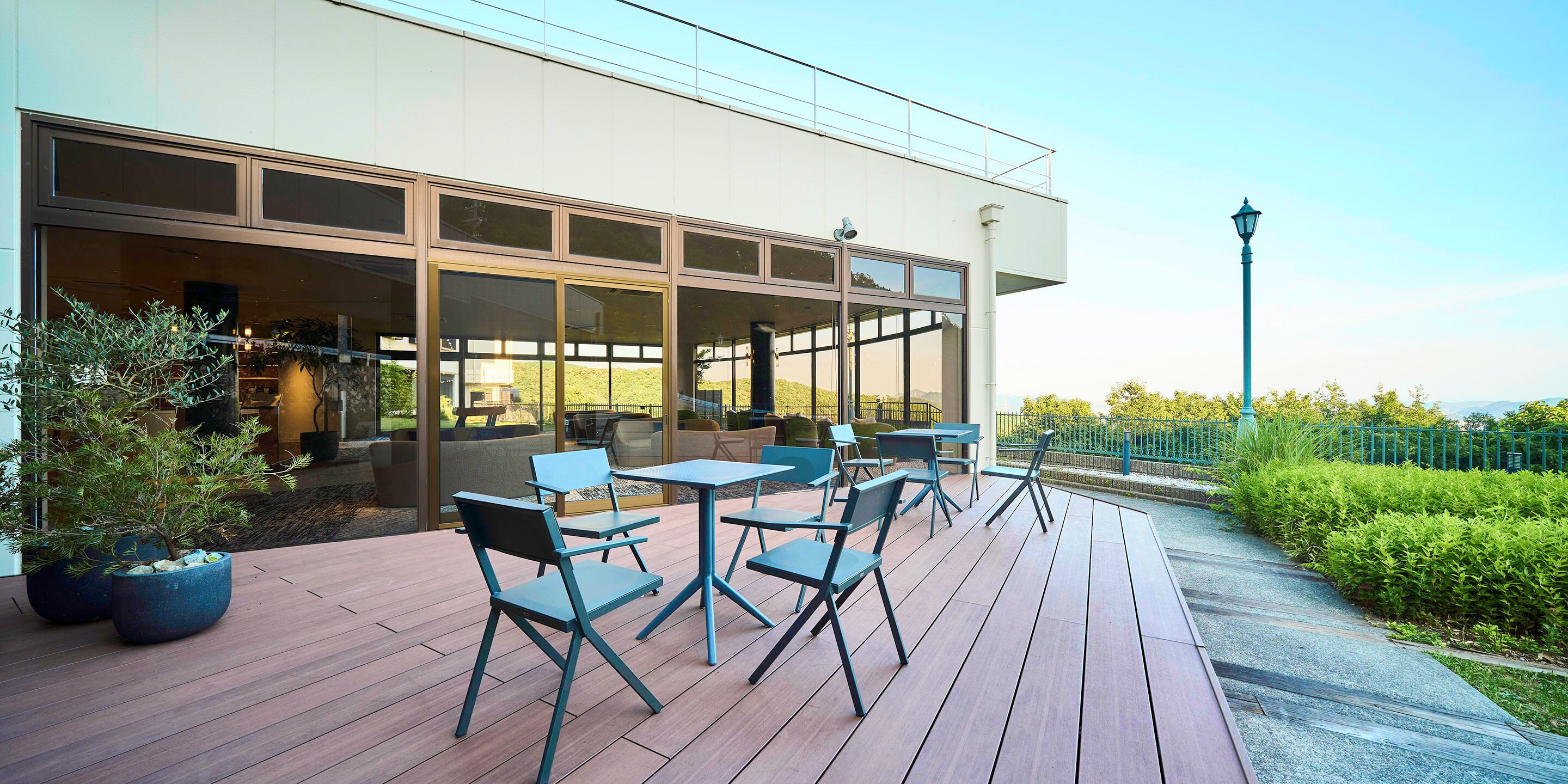 An outdoor seating area with tables and chairs, a wooden deck, and a view of the building's interior through large windows.