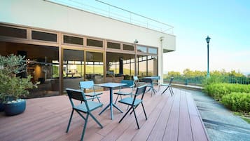 An outdoor seating area with tables and chairs, a wooden deck, and a view of the building's interior through large windows.