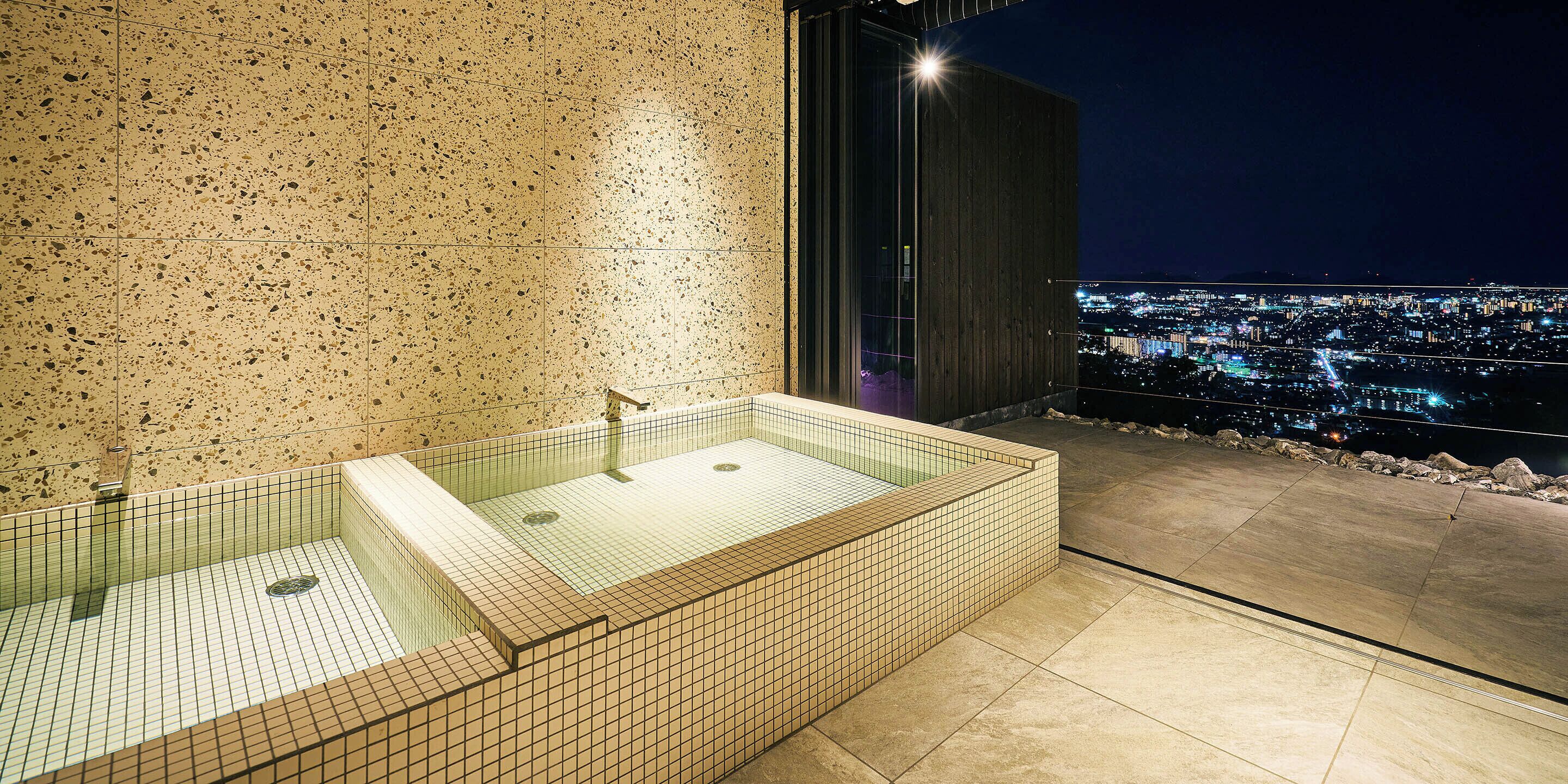 A modern bathroom with a glass-enclosed bathtub, a tiled wall, and a city view at night.