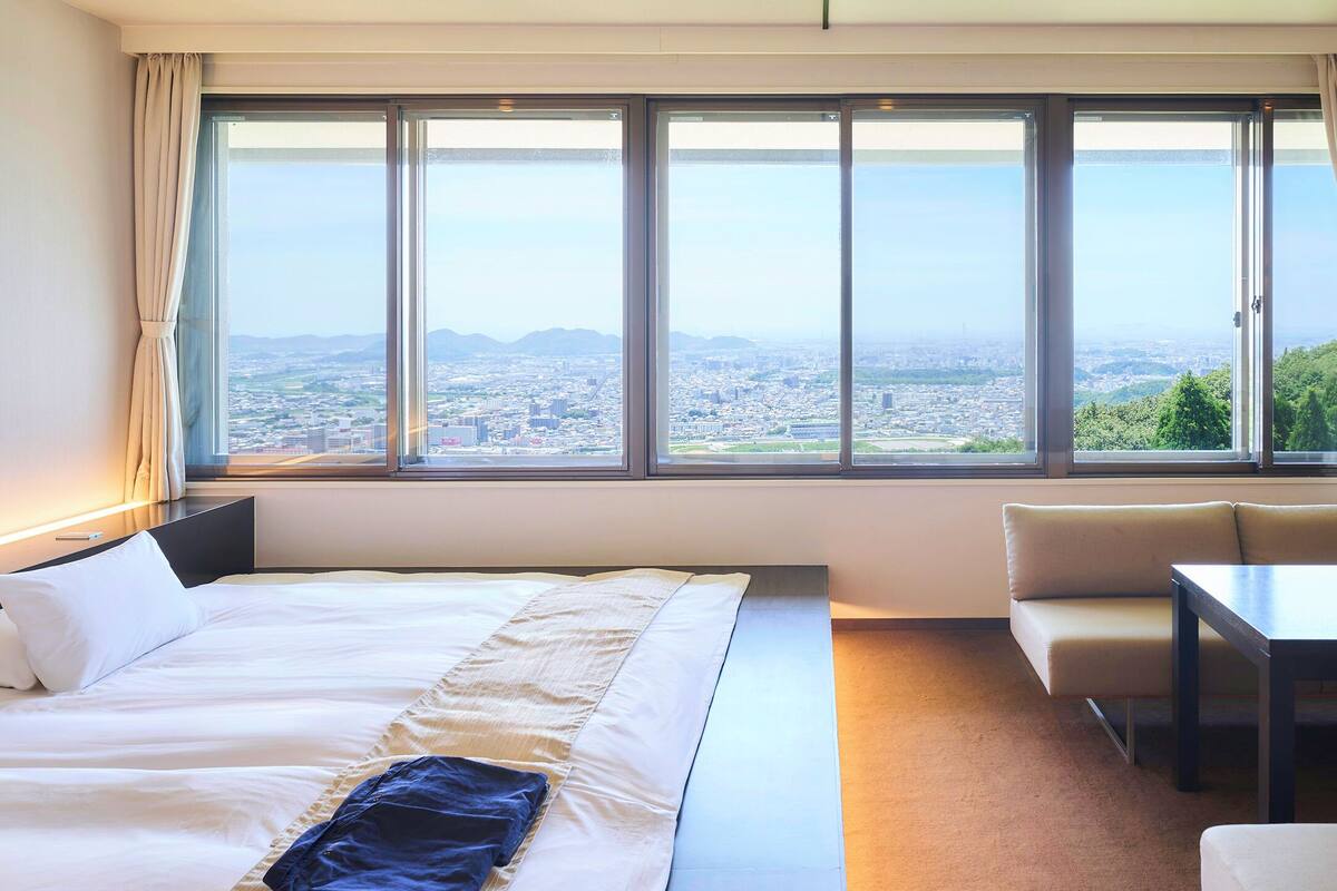 A hotel room with a large window offering a cityscape view, a bed, a sofa, and a table.