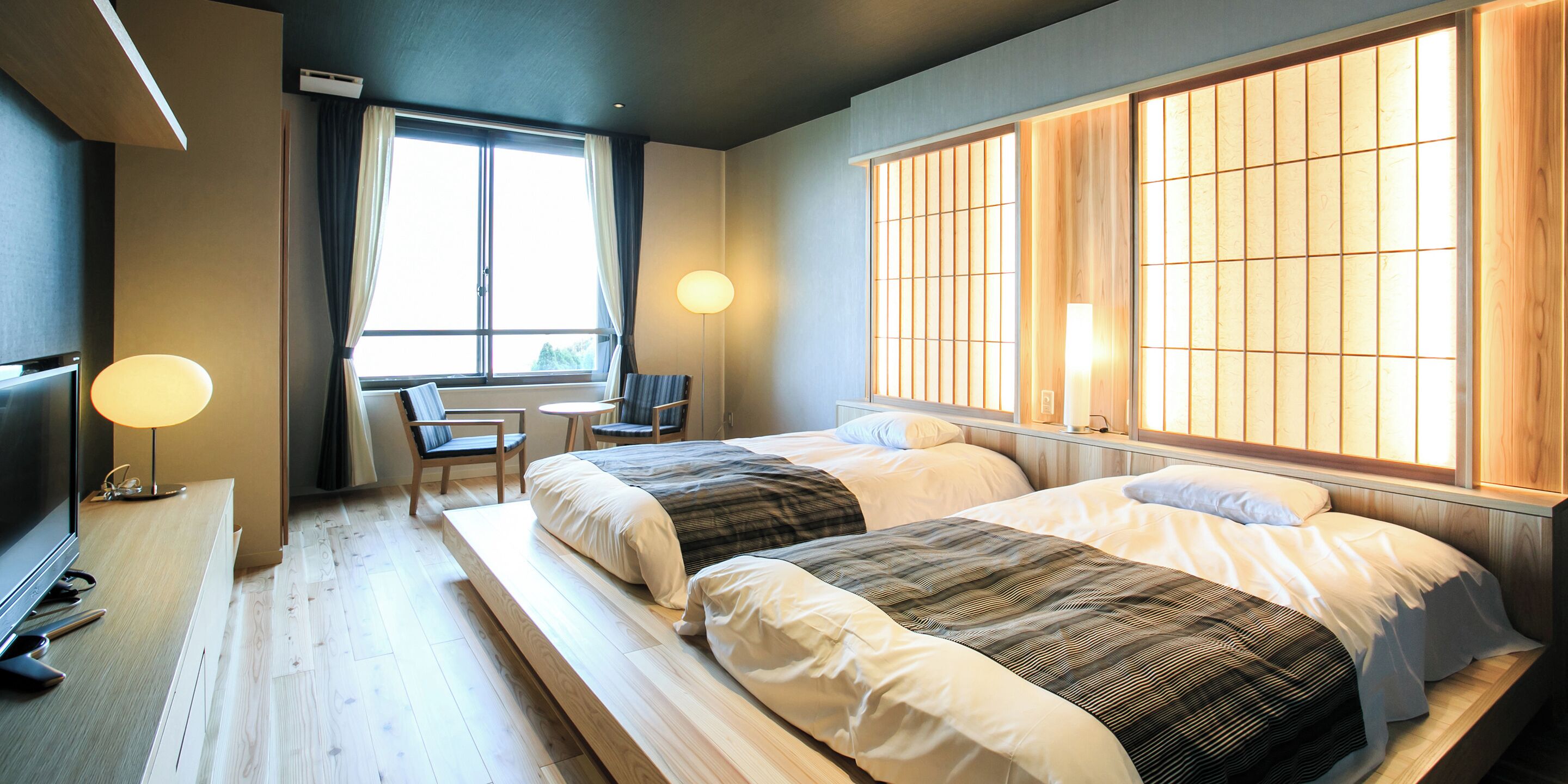 A hotel room with two beds, a desk with a chair, a TV, and large windows with wooden slats.
