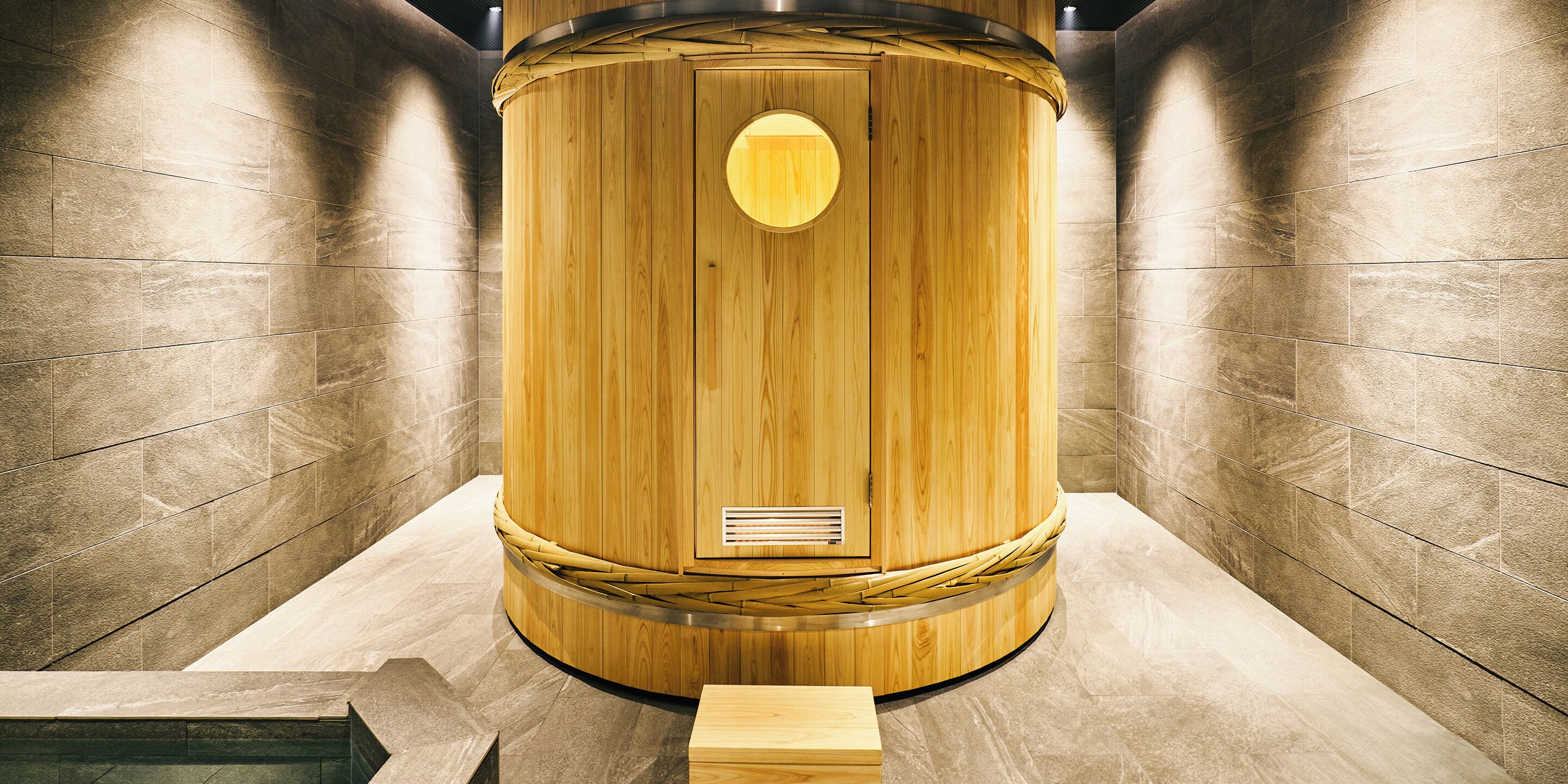 A wooden cylindrical structure with a circular window and ventilation slats, set against a tiled wall.