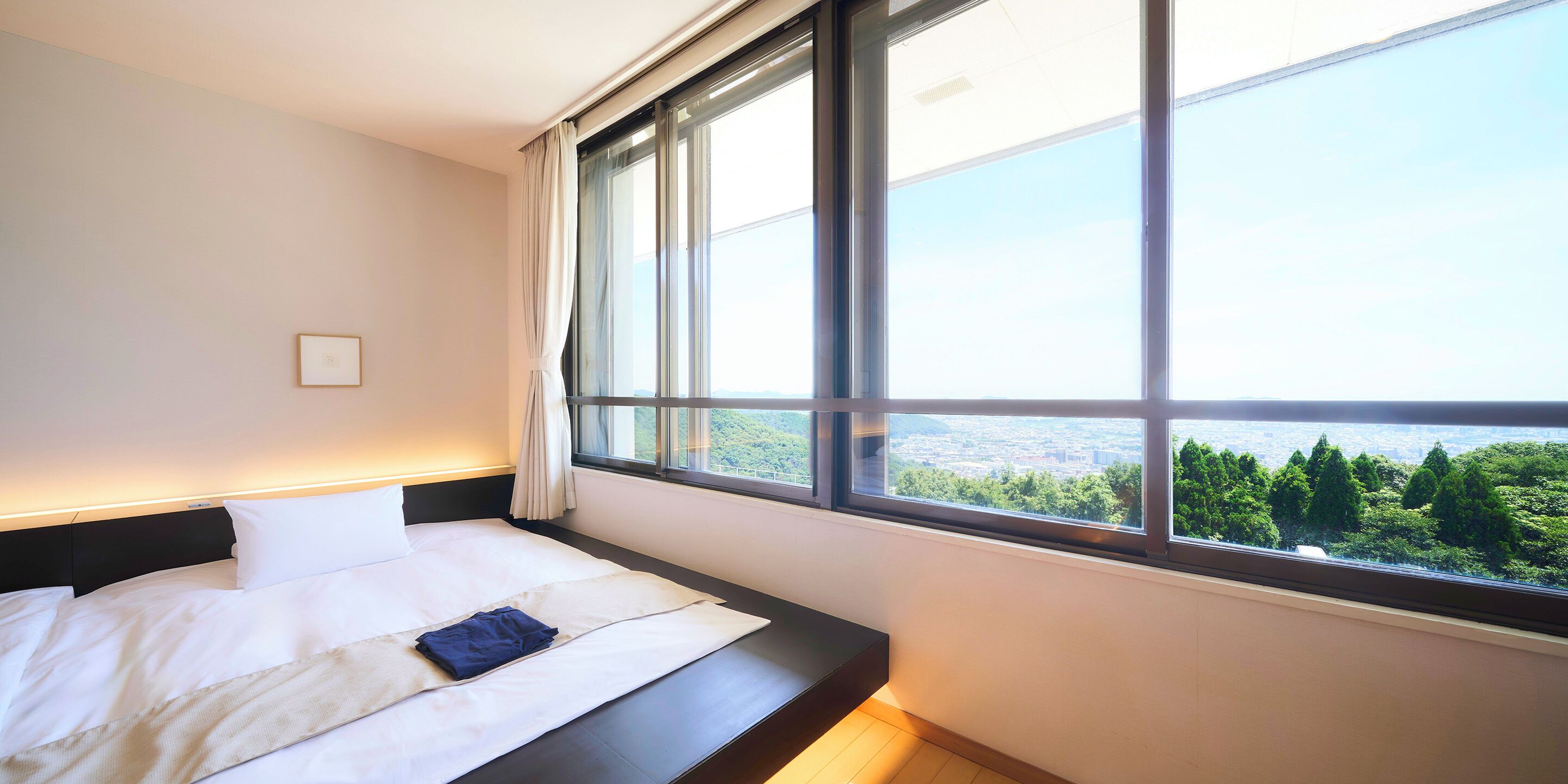A hotel room with a large window, a bed with white linens, and a view of a cityscape and greenery.