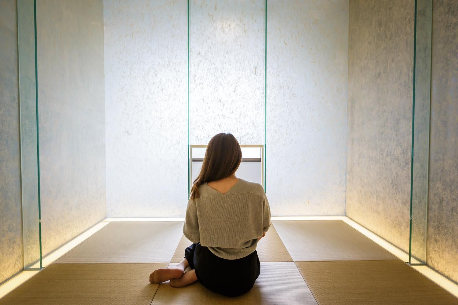 Una persona sentada sobre esteras de tatami en una habitación con diseño minimalista, ventanas amplias y una pared lisa.