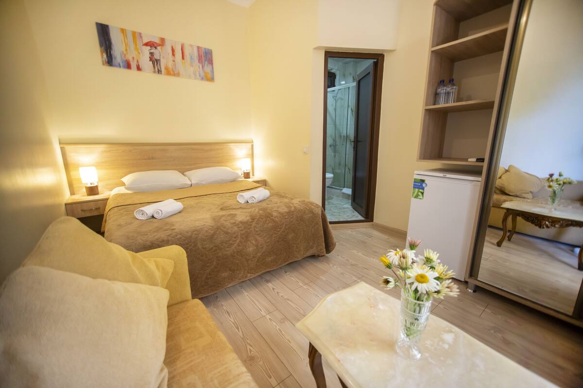 A hotel room with a bed, a sofa, a table with a vase of flowers, a mirror, and a door leading to a bathroom.