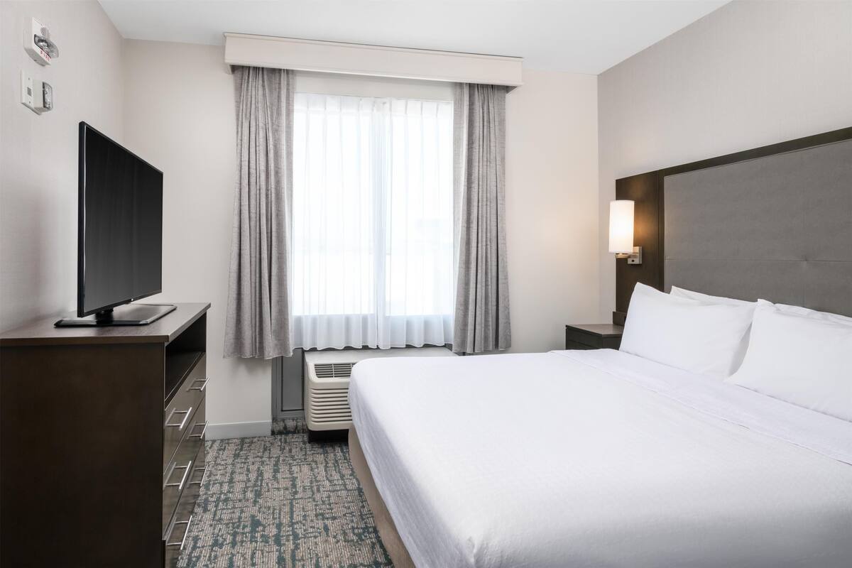 Une chambre d’hôtel avec un lit, un téléviseur posé sur un meuble, une table de chevet, une lampe et une fenêtre avec des rideaux.