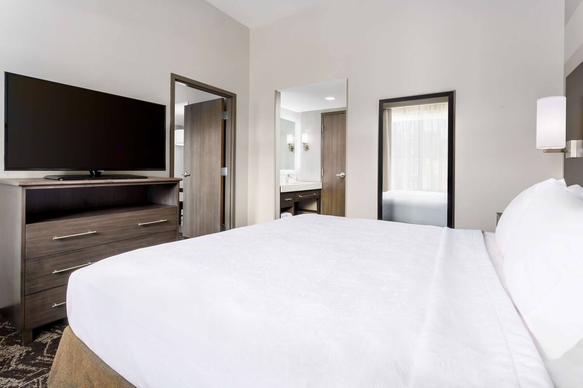 Une chambre d’hôtel avec un grand lit, un téléviseur à écran plat, une commode et une salle de bain visible à travers une porte ouverte.