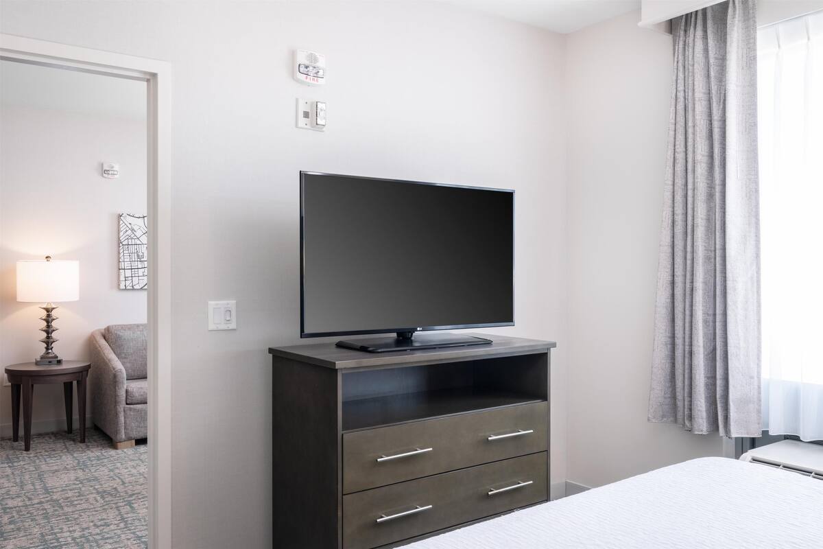 Une chambre d’hôtel équipée d’un téléviseur à écran plat posé sur un meuble en bois foncé, d’une chaise grise, d’une petite table avec une lampe et d’un lit recouvert d’une couette blanche.