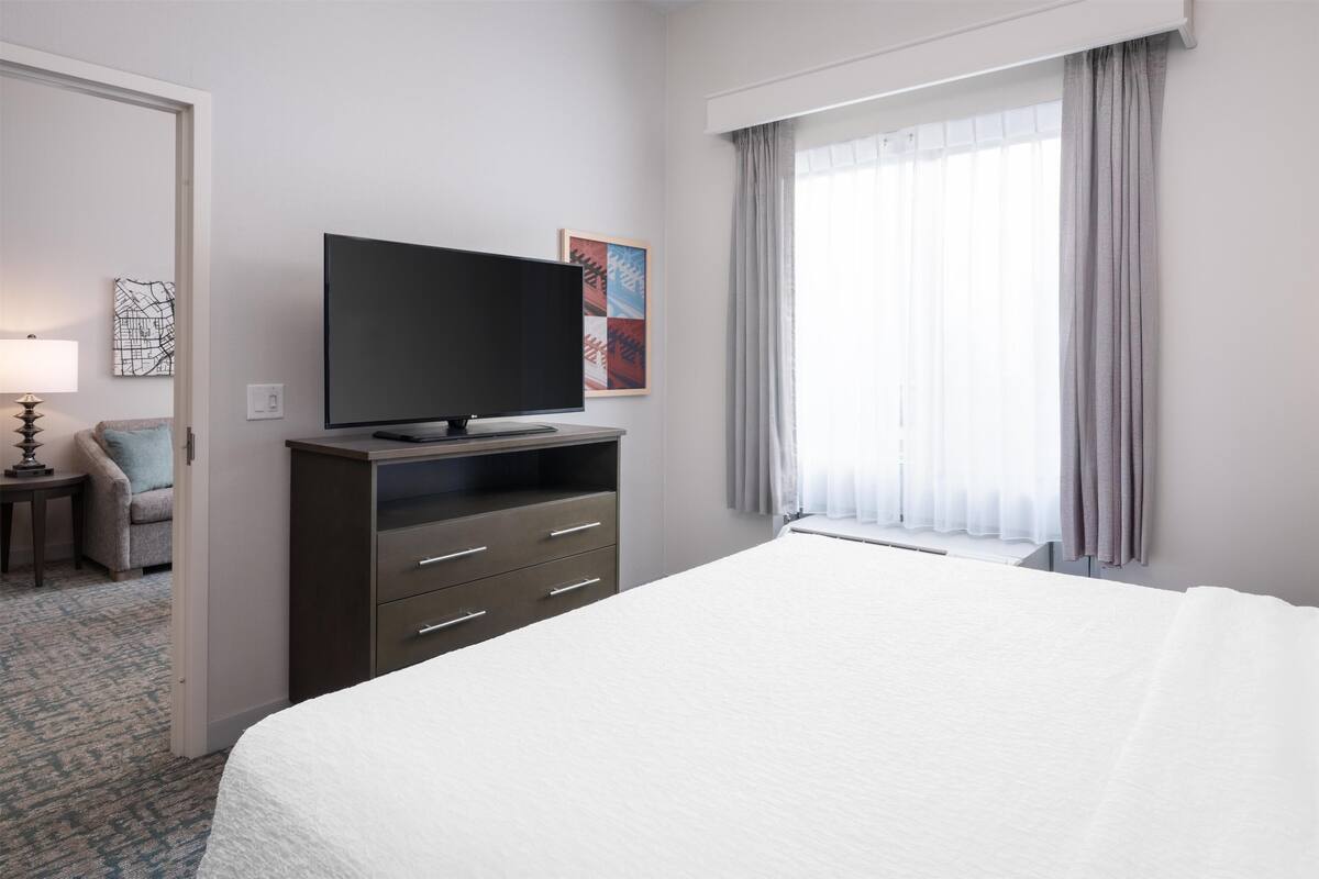 Une chambre d’hôtel avec un lit, un téléviseur posé sur un meuble, une chaise et une fenêtre avec des rideaux.