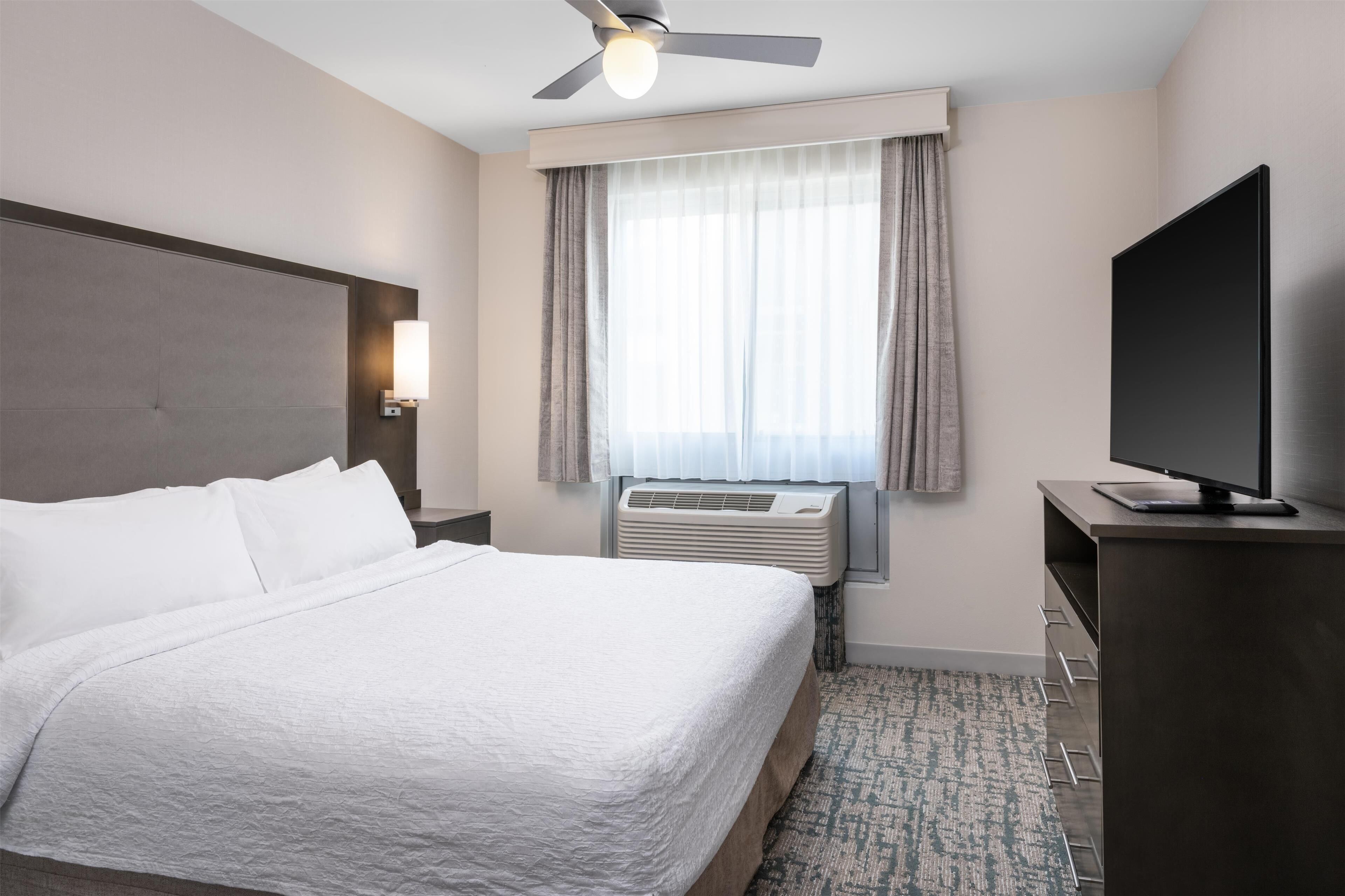 A hotel room with a bed, a flat-screen TV, a ceiling fan, an air conditioning unit, and a window with curtains.