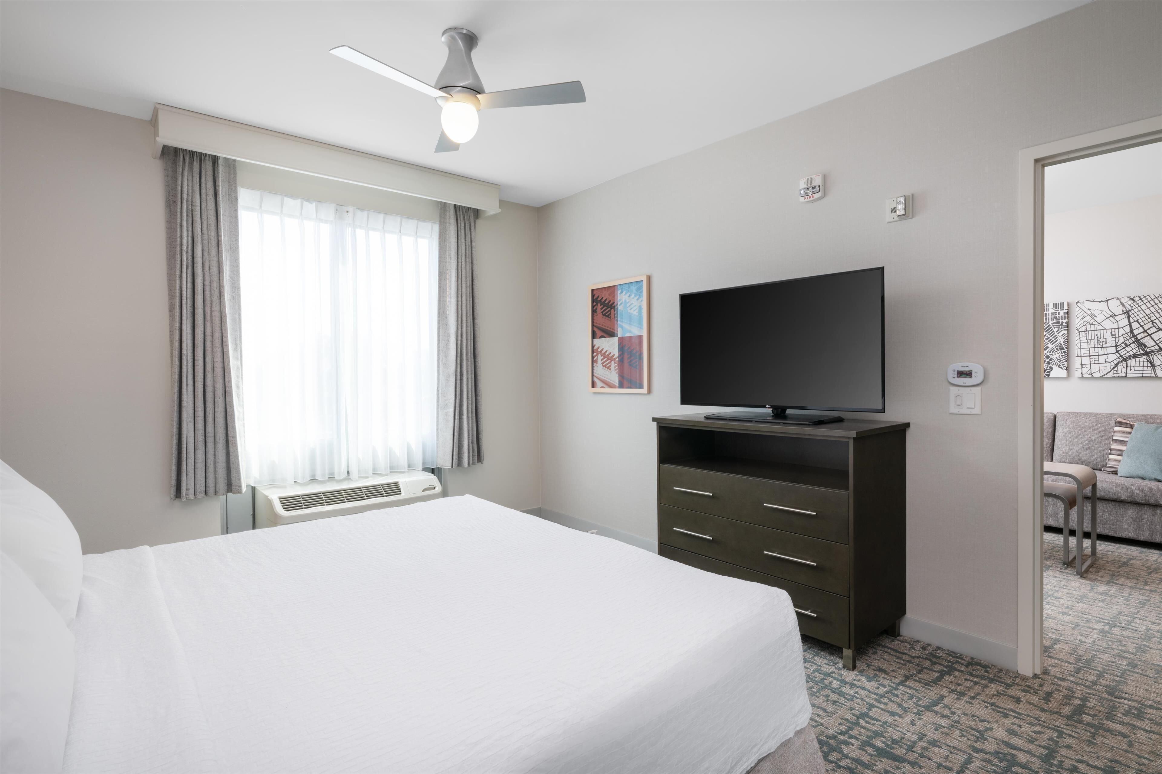 A hotel room with a bed, a flat-screen TV, a ceiling fan, and a window with curtains.