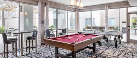 Una mesa de billar en una sala bien iluminada con ventanas amplias, una zona de bar y un área de estar informal.