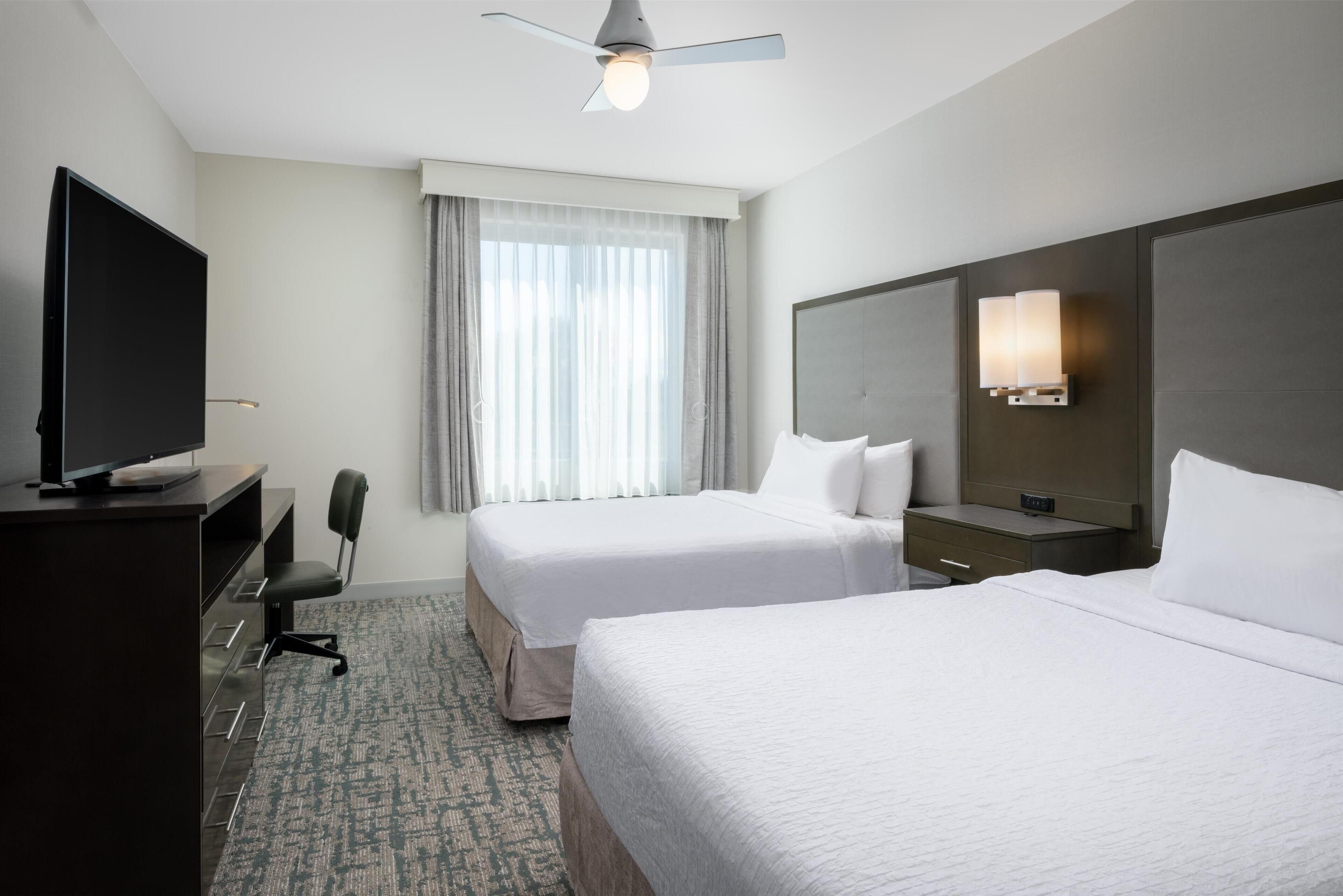 Habitación de hotel con dos camas, un escritorio con televisor de pantalla plana, una silla, un ventanal con cortinas, un ventilador de techo y una lámpara fijada a la pared.
