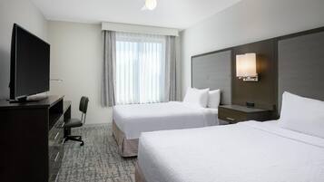 Habitación de hotel con dos camas, un escritorio con televisor de pantalla plana, una silla, un ventanal con cortinas, un ventilador de techo y una lámpara fijada a la pared.