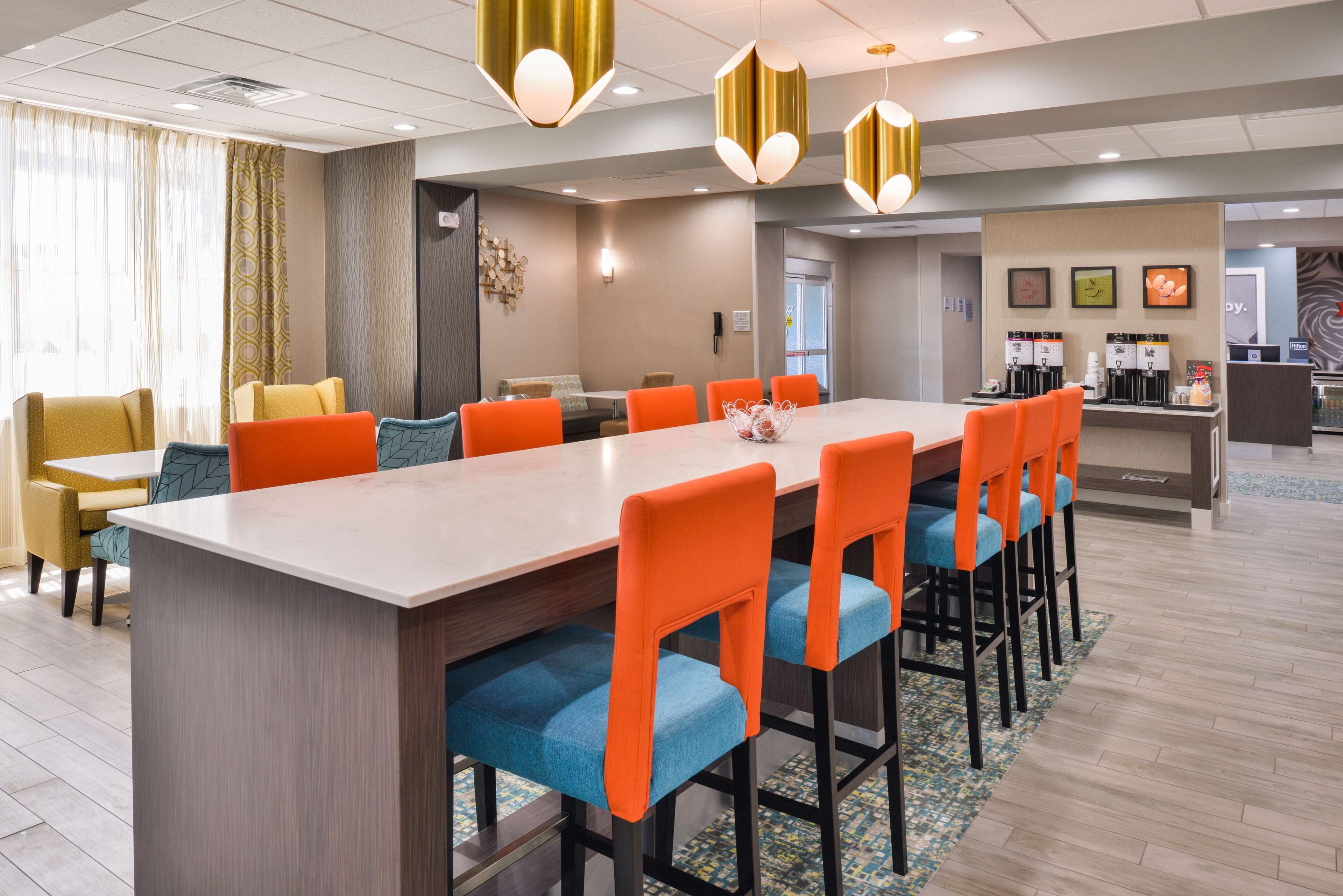 Uma sala de jantar de hotel com uma longa mesa retangular, cadeiras laranja e um balcão de bar com máquinas de café.