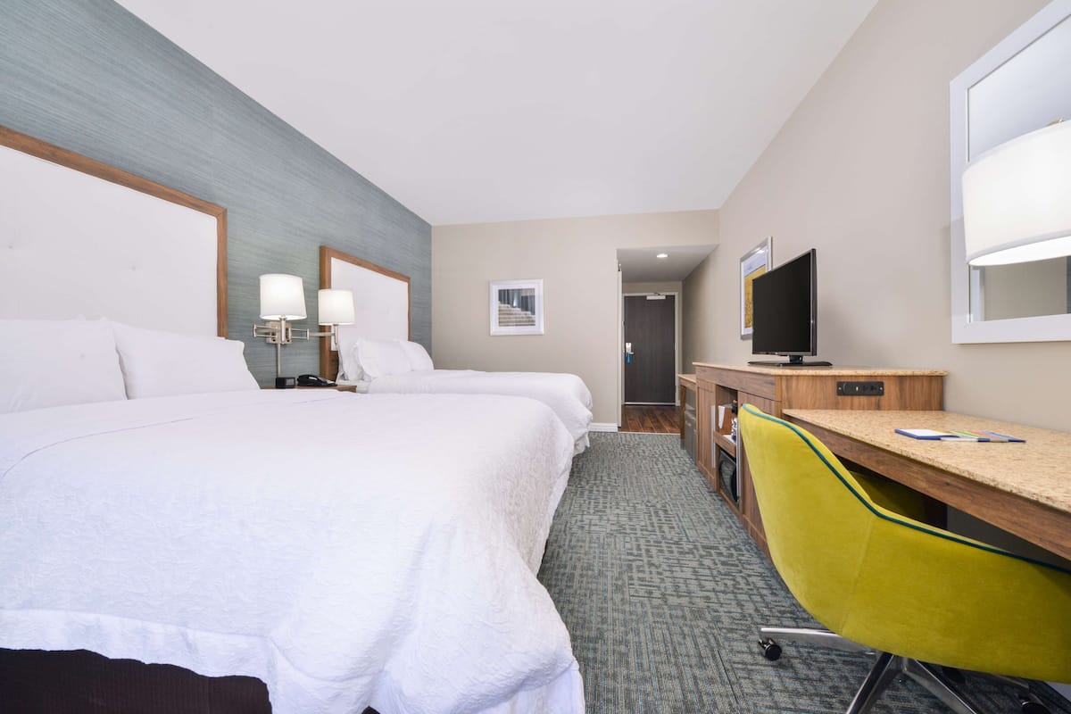 A hotel room with two beds, a desk, a chair, a television, and a bathroom visible in the background.