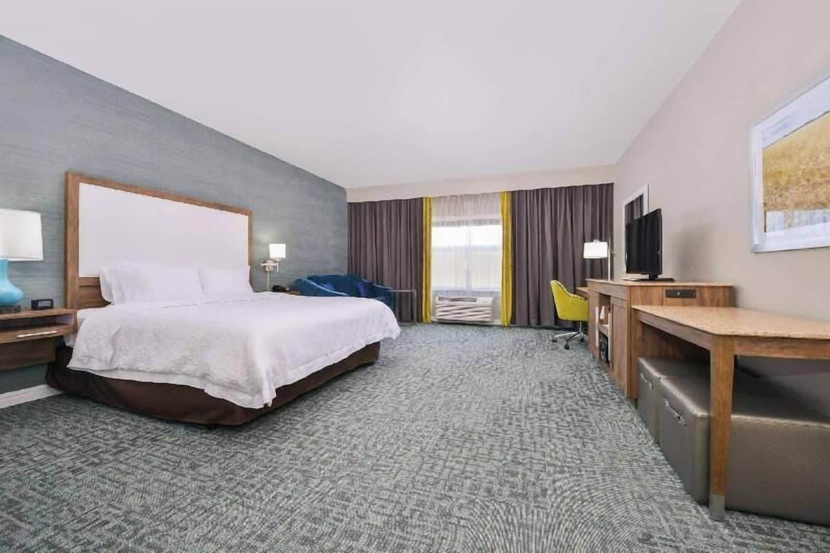 A hotel room with a bed, desk, chair, TV, and a large window with curtains.