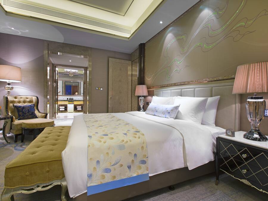 A hotel room with a large bed, a bedside table with a lamp, a chair, and a small table with a vase of flowers.
