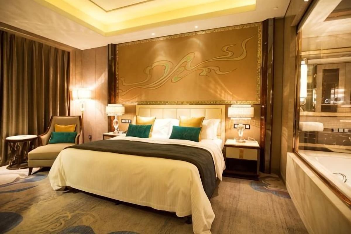 A hotel room with a large bed, two bedside tables, a headboard with a decorative panel, a sitting area with a chair, and a large window.
