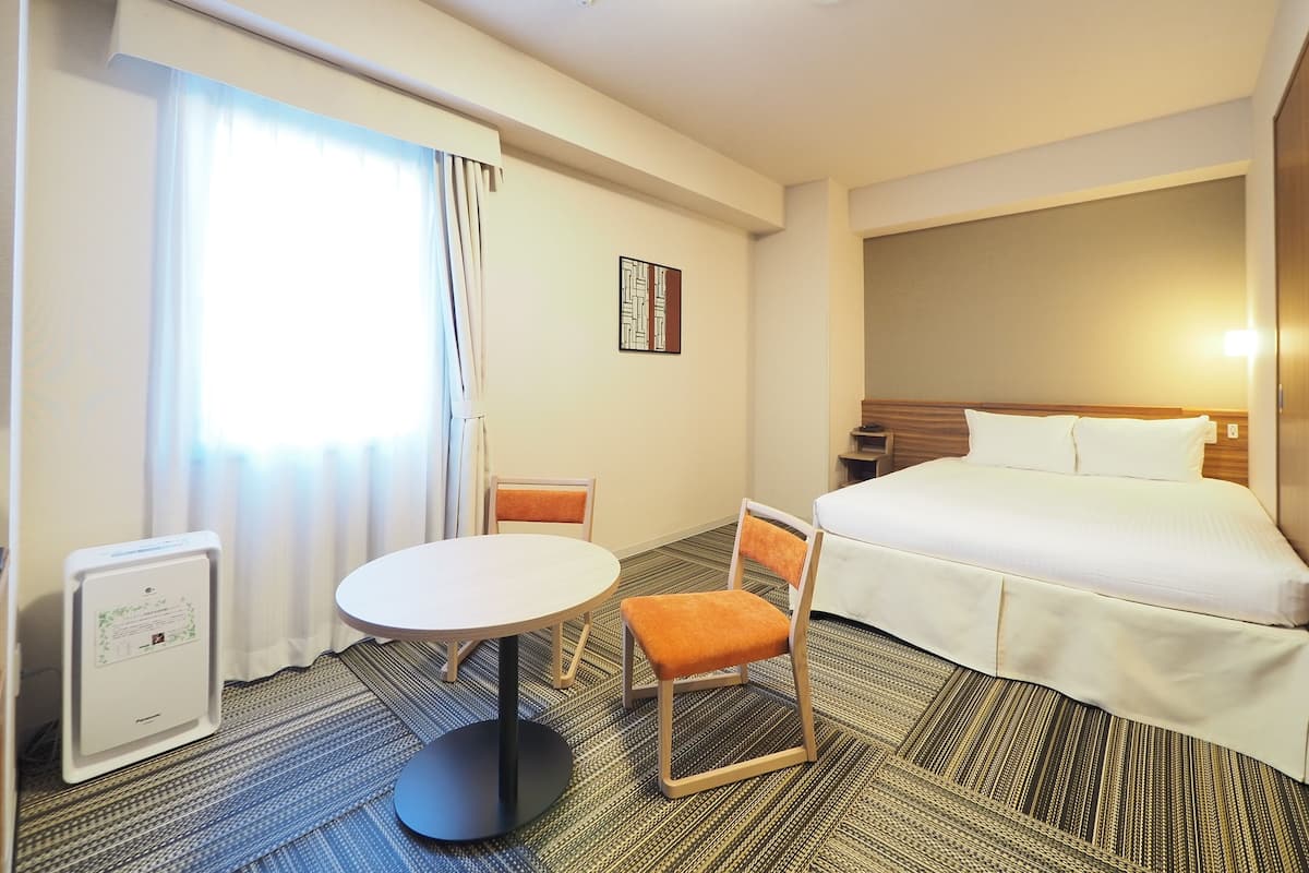 Ein Hotelzimmer mit Bett, rundem Tisch, Stuhl, kleinem Tisch mit einem Gerät und einem Fenster mit Vorhängen.