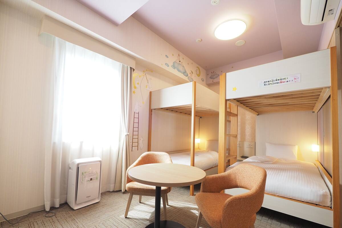 Ein Hotelzimmer mit Etagenbetten, einem runden Tisch, zwei Stühlen, einem Schreibtisch mit Lampe und einem Fenster mit Vorhängen.