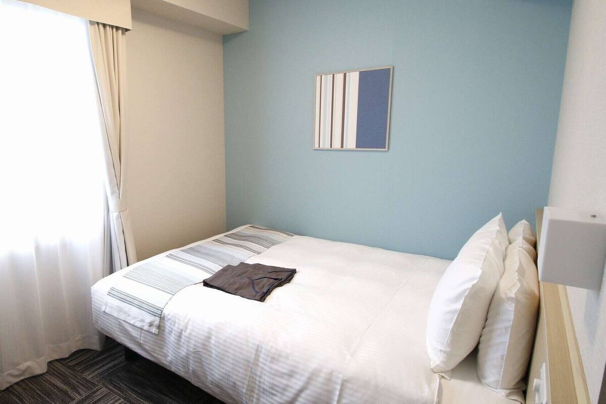 Ein Hotelzimmer mit Bett, einem Fenster mit Vorhängen, einem Wandbild und einem Nachttisch.