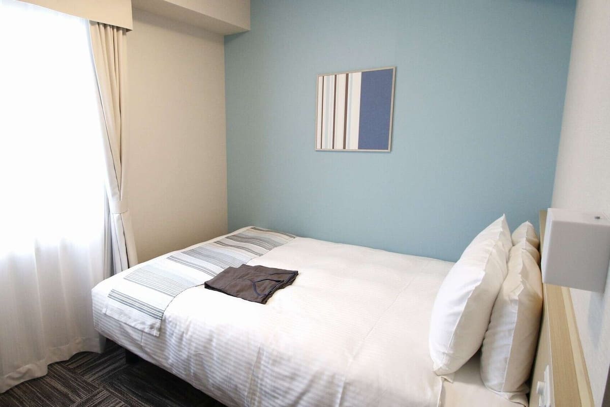 A hotel room with a bed, a window with curtains, a wall-mounted artwork, and a bedside table.