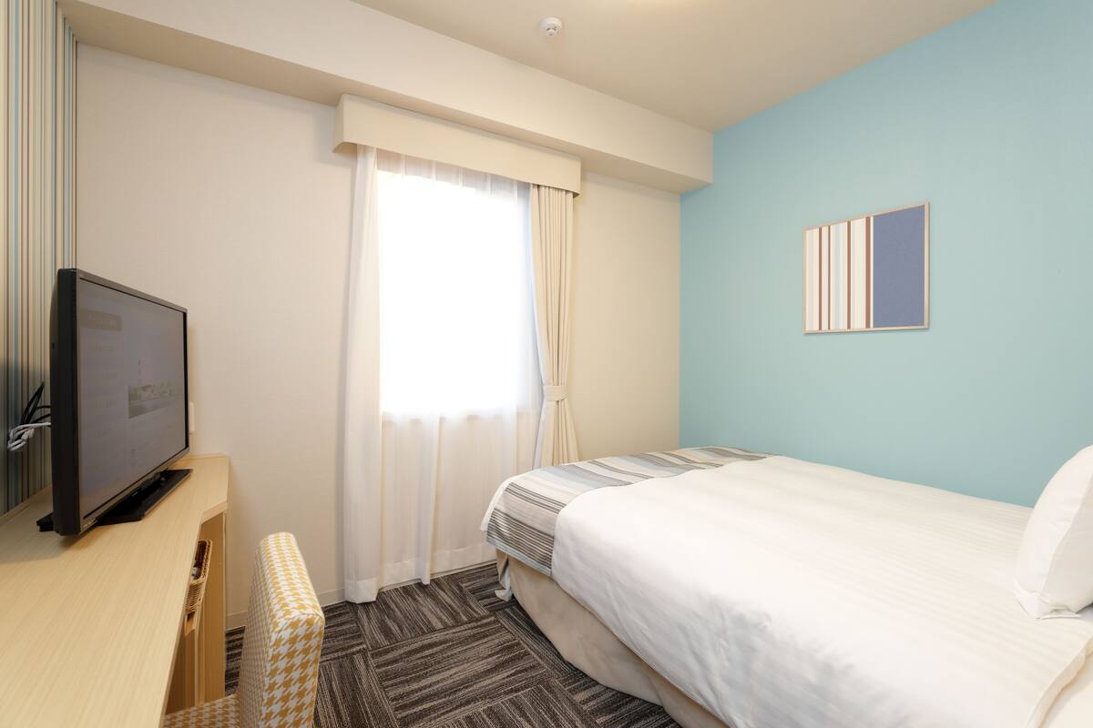 Ein Hotelzimmer mit Bett, Fernseher, Fenster mit Vorhängen, Wandbild und Teppichboden.