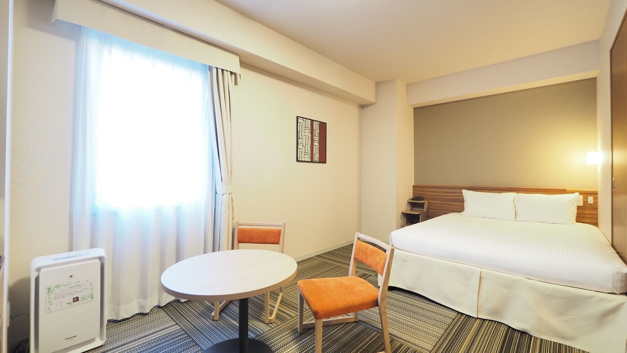 A hotel room with a bed, a round table, a chair, a radiator, and a window with curtains.